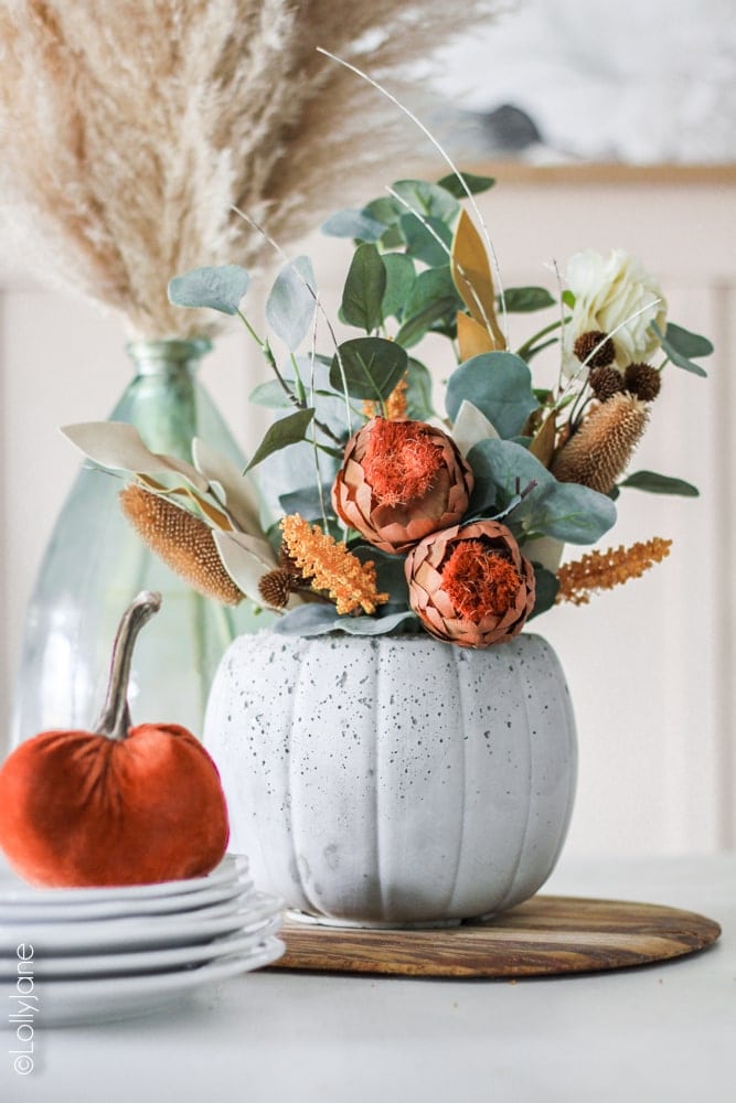 DIY Concrete Pumpkin Planter... perfect for fall and SURPRISE: the back has a jack-o-lantern face on it, also perfect for Halloween! Made from a $1 treat bucket, this is a CHEAP yet CHIC project anyone can make! #diy #halloweendecor #falldecor #fallplanter #pumpkinplanter #diypumpkin #pumpkinfloralarrangement #fallfloralarrangement #fallflowers #pumpkinplanter
